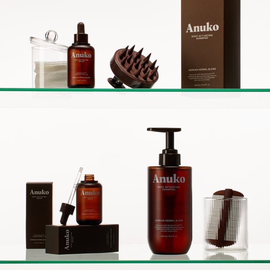 Anuko's range on glass shelves: Root Activating Serum and Shampoo in amber bottles, a scalp brush and cotton pads